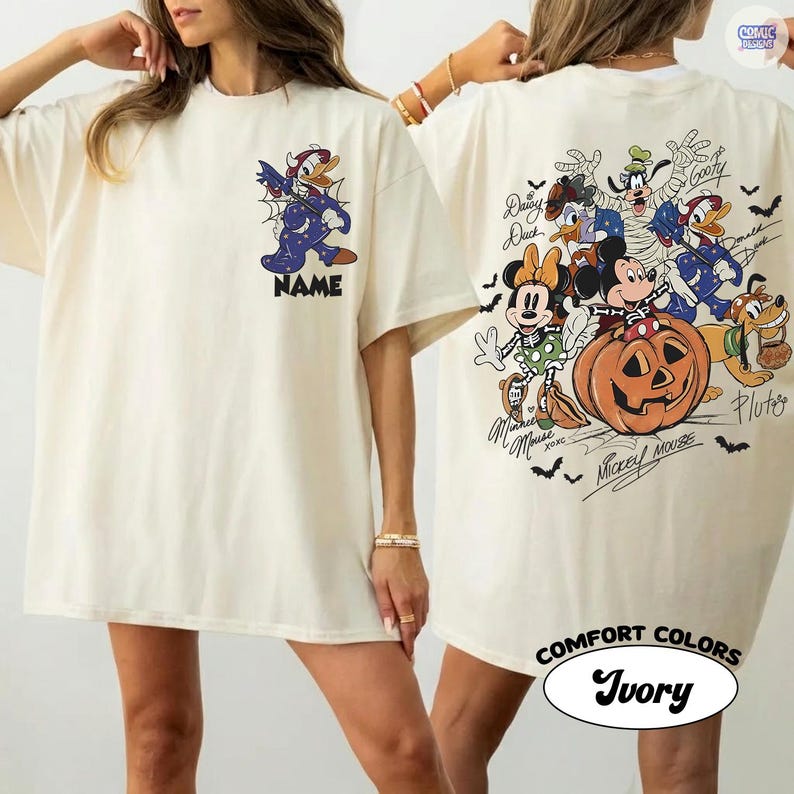 May include: An ivory t-shirt featuring a small graphic of Donald Duck in a costume on the front chest with the word NAME below it. The back displays a large Halloween-themed illustration of Mickey Mouse, Minnie Mouse, Goofy, Donald Duck, and Pluto.