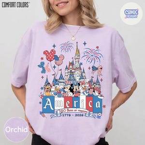 May include: A light purple short-sleeve t-shirt featuring a graphic of a castle, fireworks, and cartoon characters wearing patriotic red, white, and blue clothing. The text reads America, 250 Years of Freedom, and 1776-2026 in a banner below.