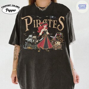 May include: Black Comfort Colors t-shirt with a pirate-themed graphic. The design features the word "Pirates" in a vintage font, a cartoon pirate character, a treasure chest, and a pirate ship. The shirt has the "Comic Designs" and "Pepper" logos.