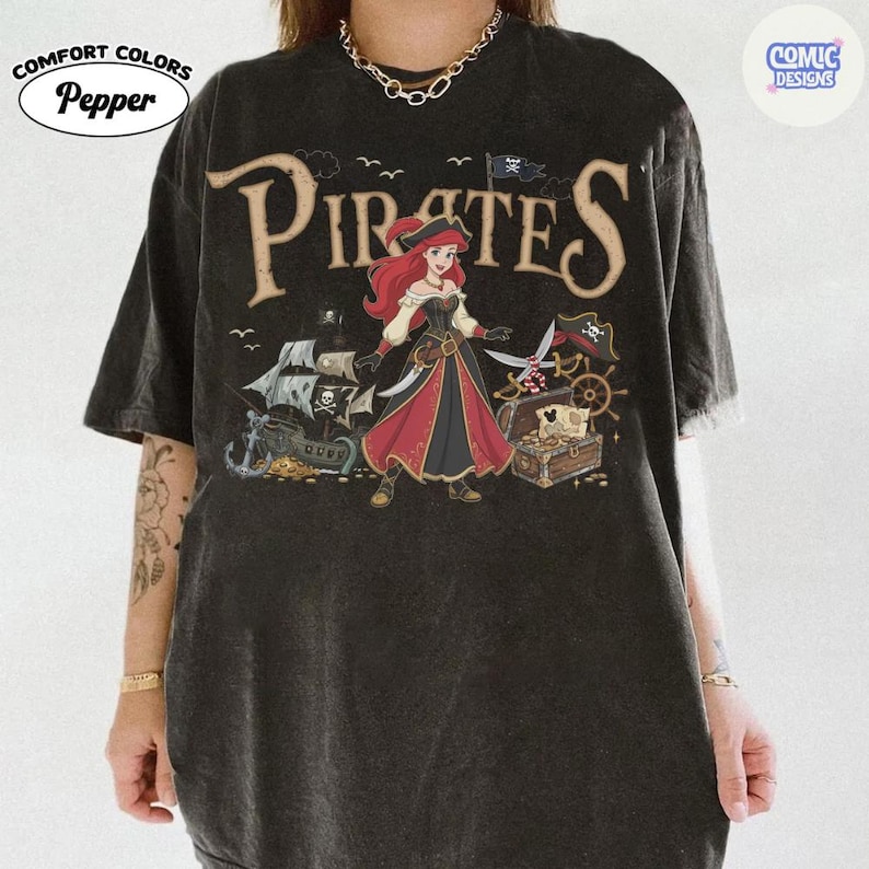 May include: Black Comfort Colors t-shirt with a pirate-themed graphic. The design features the word "Pirates" in a vintage font, a cartoon pirate character, a treasure chest, and a pirate ship. The shirt has the "Comic Designs" and "Pepper" logos.