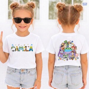 May include: A white short-sleeved t-shirt featuring the name CAMILA on the front in colorful, character-themed letters. The back of the shirt displays a graphic print of various animated toy characters gathered around a purple backpack.