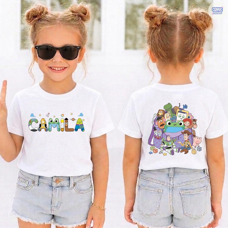 May include: A white short-sleeved t-shirt featuring the name CAMILA on the front in colorful, character-themed letters. The back of the shirt displays a graphic print of various animated toy characters gathered around a purple backpack.