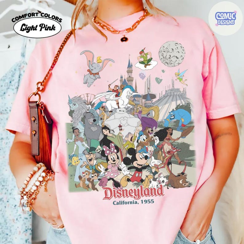 Puede incluir: Camiseta rosa claro Comfort Colors con un dise&ntilde;o de Disneyland. El dise&ntilde;o incluye varios personajes de Disney como Mickey y Minnie Mouse, Dumbo y Peter Pan. Tambi&eacute;n se puede ver el texto "Disneyland California, 1955".