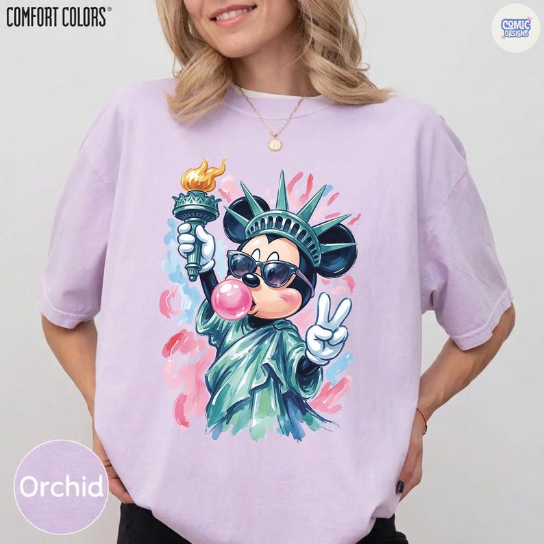 May include: A light purple short-sleeve t-shirt featuring a graphic of a cartoon mouse dressed as the Statue of Liberty, wearing sunglasses, holding a torch, and blowing a pink bubblegum bubble while making a peace sign with one hand.