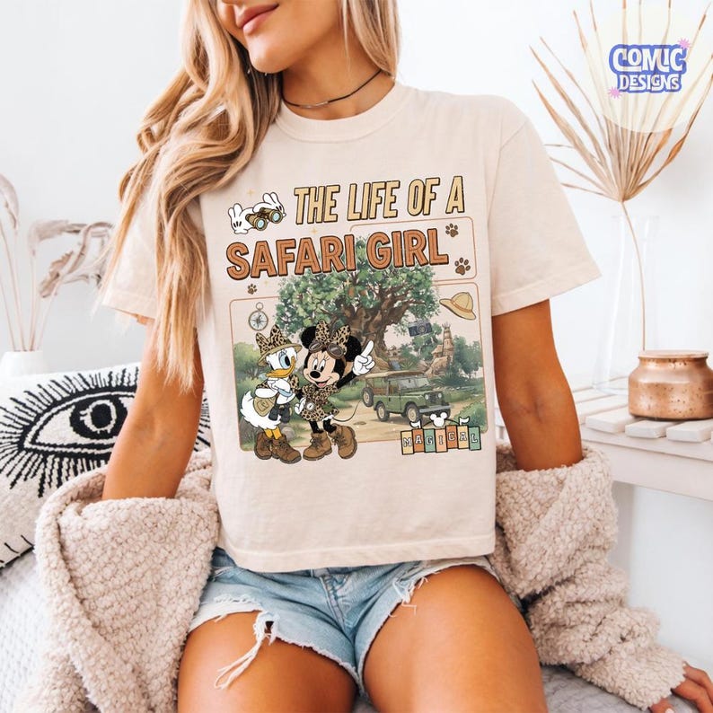 May include: Beige t-shirt with the text "THE LIFE OF A SAFARI GIRL" and a graphic of Minnie and Daisy Duck in safari outfits. The graphic includes a safari jeep, a tree, and a hat. The shirt has short sleeves.