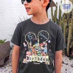 May include: Dark gray t-shirt featuring a colorful graphic of cartoon characters, the number 5, and the name "Joseph" in a heart-shaped design. The text "Birthday Squad" is also displayed. The shirt is worn by a young person.