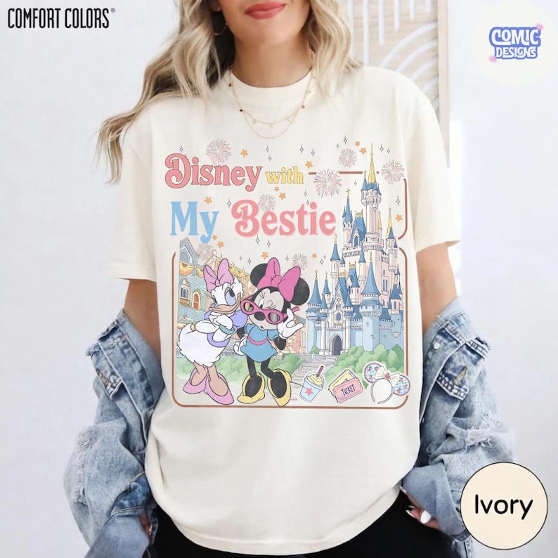 K&ouml;nnte beinhalten: Elfenbeinfarbenes T-Shirt mit dem Text "Disney with My Bestie" und einer Grafik mit Daisy Duck, Minnie Mouse und einem Schloss. Das Shirt ist von der Marke Comfort Colors und hat ein Comic Designs Logo.