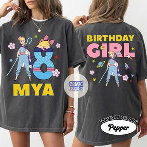 Puede incluir: Camiseta gris oscuro con un dise&ntilde;o de personaje de dibujos animados, el n&uacute;mero 8 y el nombre "MYA" en amarillo. La parte trasera de la camiseta dice "BIRTHDAY GIRL". La camiseta est&aacute; etiquetada como "COMFORT COLORS Pepper".