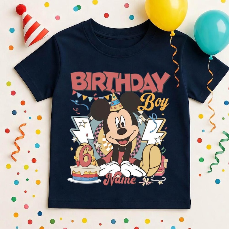 May include: Navy blue t-shirt with a colorful graphic featuring Mickey Mouse, the text "BIRTHDAY Boy", and the number "6". The design includes a birthday cake and gift boxes. Party decorations like balloons and confetti are in the background.