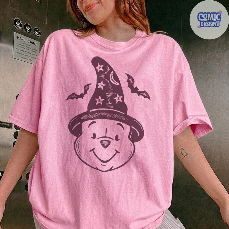 May include: Pink t-shirt featuring a cartoon bear wearing a witch hat with stars and a crescent moon. Two bat silhouettes flank the hat. The shirt has a relaxed fit and short sleeves, perfect for casual wear.