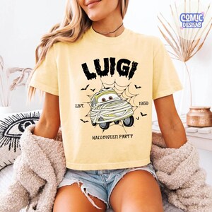 May include: A pale yellow short-sleeve t-shirt featuring a graphic of a cartoon car wrapped in bandages like a mummy. The text reads LUIGI, EST. 1959, and HALLOWEEN PARTY, surrounded by a spider web and small black bats printed in black ink.