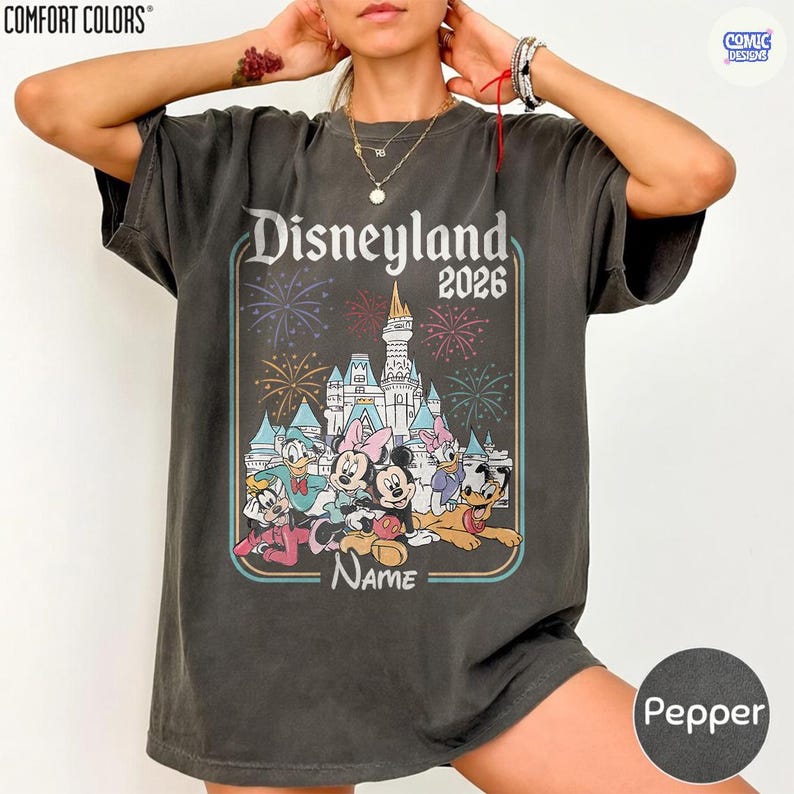 May include: Pepper-colored t-shirt featuring a Disneyland 2026 graphic. The design includes a castle, fireworks, and Disney characters like Mickey and Minnie Mouse. The shirt has a vintage, worn look.