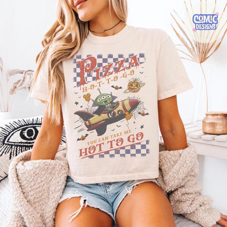 May include: A cream-colored short-sleeve t-shirt featuring a graphic of a green alien in a rocket ship. The design includes a checkered pattern, spider webs, pumpkins, and the text "Pizza H-O-T-T-O-G-O" and "You can take me hot to go".