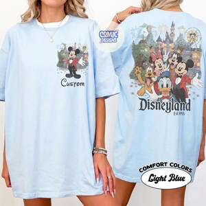 May include: A light blue t-shirt featuring a small graphic of Mickey Mouse on the front with the word Custom below it. The back displays a large illustration of Mickey, Minnie, Donald, and Pluto in front of a castle with the text Disneyland Est. 1955.