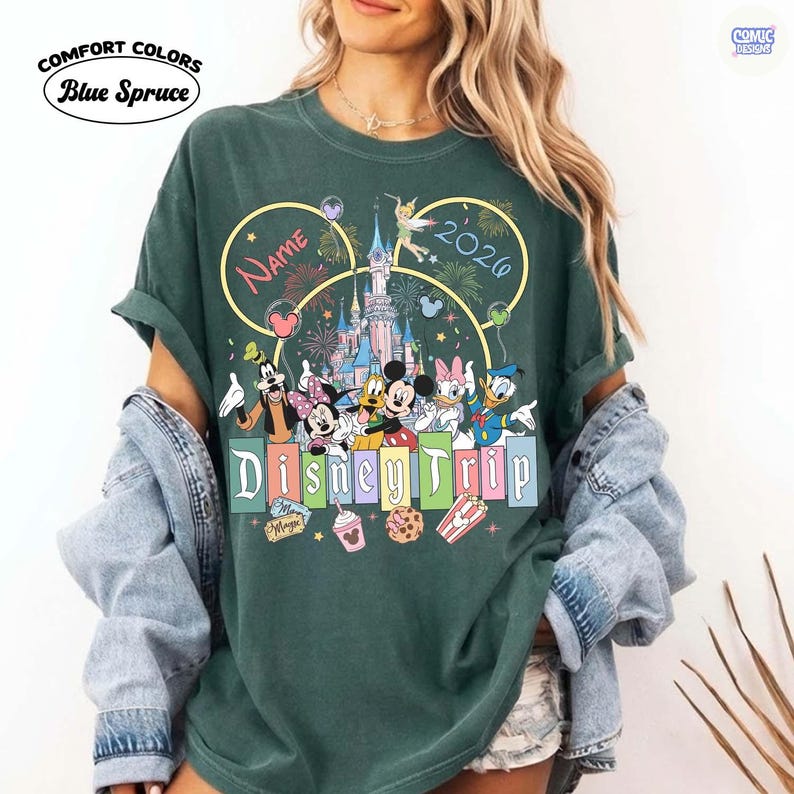 May include: Green Comfort Colors t-shirt with a Disney Trip design. The design features Mickey Mouse, Minnie Mouse, Donald Duck, and other Disney characters in front of a castle. The shirt includes the text "Name" and "2026".