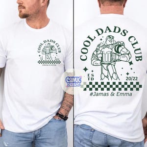 May include: White t-shirt with a green graphic of Buzz Lightyear holding a beer mug, with the text "COOL DADS CLUB". The shirt also features the text "ES TD 20/22" and "#Jamas & Emma".