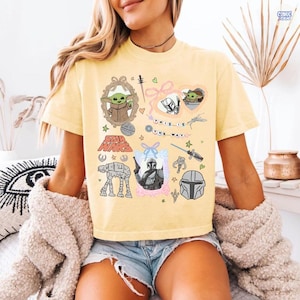 Puede incluir: Camiseta amarilla clara con gr&aacute;ficos tem&aacute;ticos de Star Wars. El dise&ntilde;o incluye im&aacute;genes de Grogu, el Mandaloriano y el texto "THIS IS THE WAY". La camiseta tiene cuello redondo y mangas cortas.