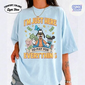May include: Light blue Comfort Colors t-shirt with a graphic of Goofy holding money, surrounded by dollar bills and popcorn. Text reads "I'M JUST HERE TO PAY FOR EVERYTHING". The shirt has a relaxed fit.