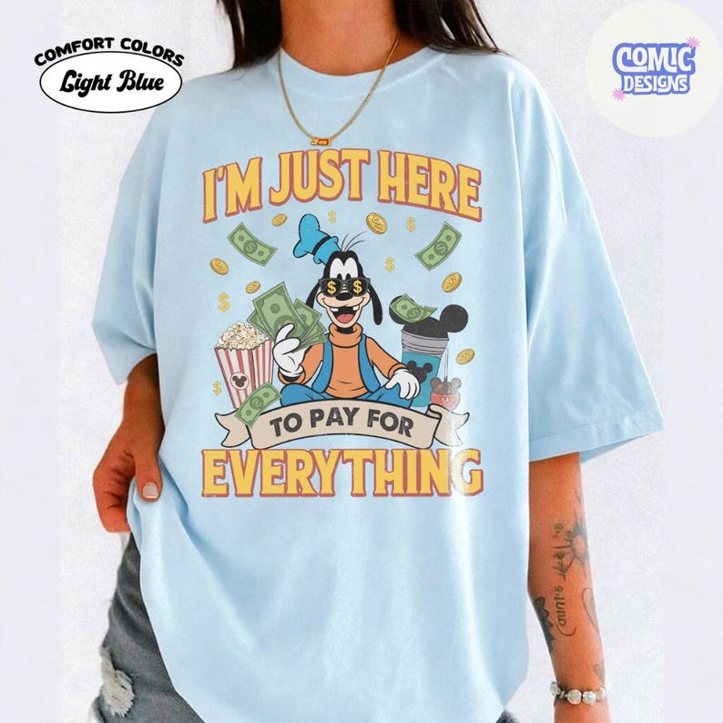 May include: Light blue Comfort Colors t-shirt with a graphic of Goofy holding money, surrounded by dollar bills and popcorn. Text reads "I'M JUST HERE TO PAY FOR EVERYTHING". The shirt has a relaxed fit.
