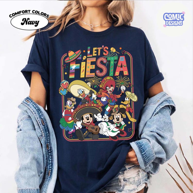 May include: Navy blue t-shirt with a colorful "Let's Fiesta" graphic featuring cartoon characters in festive attire. The design includes a sombrero, fireworks, and musical instruments. The shirt is labeled "Comfort Colors."
