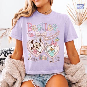 May include: A light purple t-shirt featuring the word "Besties" in pastel colors. The shirt has a graphic of Minnie Mouse and Daisy Duck in heart-shaped lockets. The design includes bows, chains, and the text "Minnie Mouse xoxo" and "Daisy Duck".