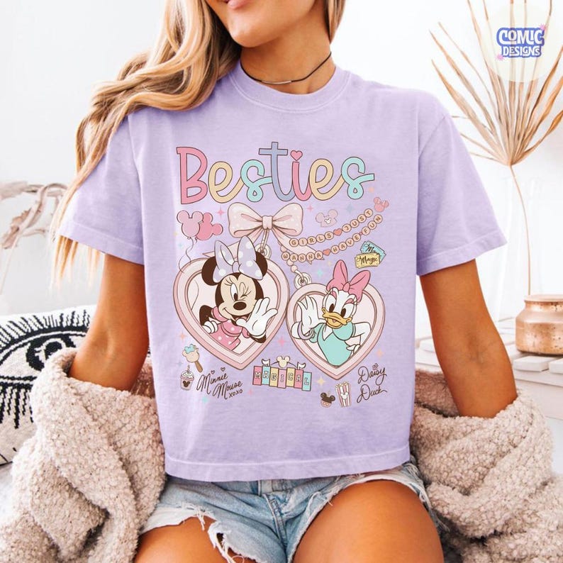May include: A light purple t-shirt featuring the word "Besties" in pastel colors. The shirt has a graphic of Minnie Mouse and Daisy Duck in heart-shaped lockets. The design includes bows, chains, and the text "Minnie Mouse xoxo" and "Daisy Duck".