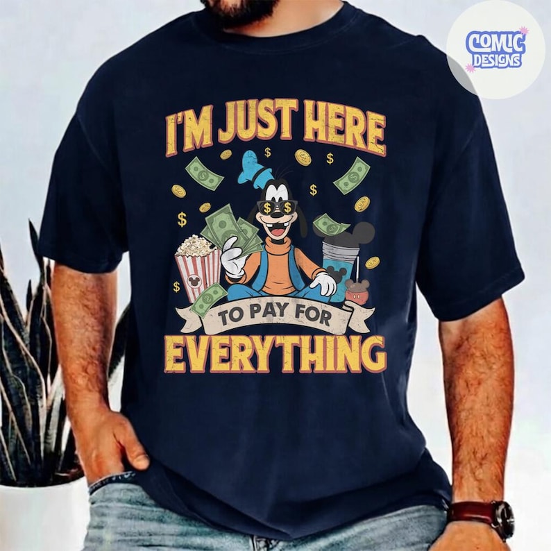 May include: Navy blue t-shirt with a graphic of Goofy holding money and popcorn, with the text "I'M JUST HERE TO PAY FOR EVERYTHING". The design includes dollar signs and a Mickey Mouse-themed drink.