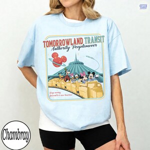 Puede incluir: Camiseta azul claro con un dise&ntilde;o de Tomorrowland Transit Authority Peoplemover. El gr&aacute;fico incluye a Mickey Mouse, Minnie Mouse, Goofy, Donald Duck y otros personajes en un tren, con el texto "Keep moving forward to a new tomorrow."