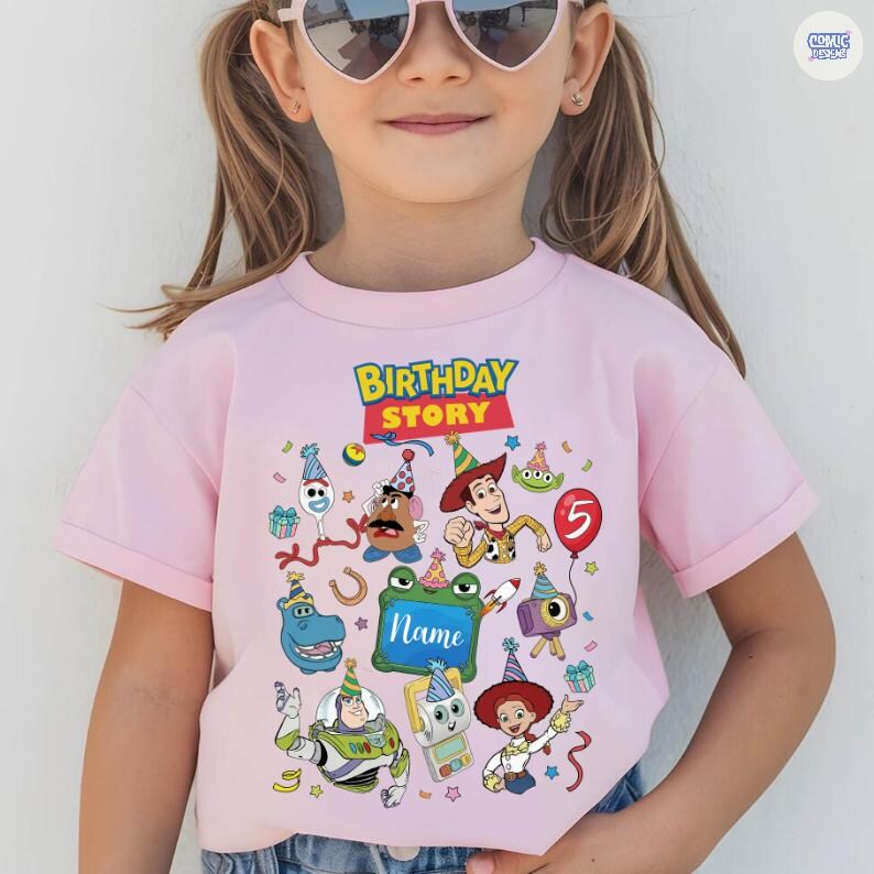 May include: A light pink t-shirt featuring a colorful graphic of various animated characters wearing party hats. The text "BIRTHDAY STORY" is printed at the top, and a central rectangular frame contains the word "Name" in blue script.