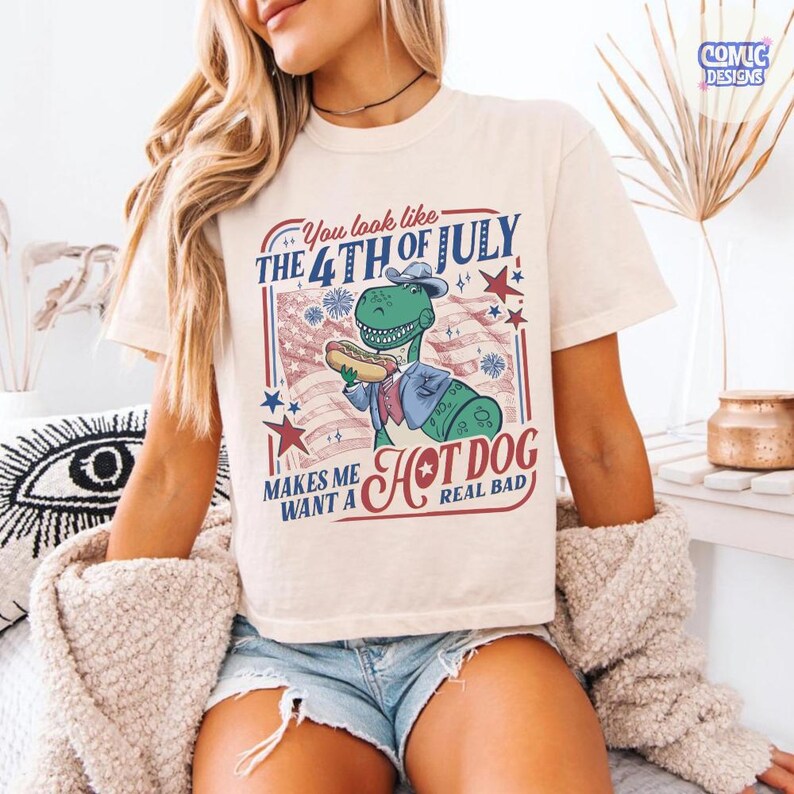 May include: Cream-colored t-shirt featuring a graphic of a dinosaur holding a hot dog, with text that reads "You look like the 4th of July makes me want a hot dog real bad." The design includes red, white, and blue elements.