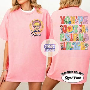 Op de afbeelding: Een lichtroze t-shirt met een afbeelding van Bo Peep in een gele cirkel en de tekst Name op de voorzijde. De achterkant toont de tekst YOU'VE GOT A FRIEND IN ME met kleurrijke illustraties van diverse Toy Story personages.
