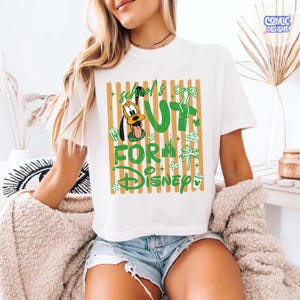 May include: White t-shirt with a green and orange graphic that says "School's Out For Disney" featuring Pluto. The design includes a palm tree, castle, and other Disney-themed elements. The shirt is a casual, short-sleeved style.