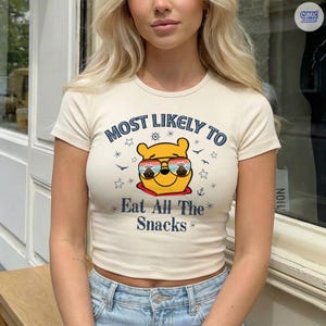 May include: Cream-colored, short-sleeved crop top with the text "MOST LIKELY TO Eat All The Snacks" in blue. The graphic features a yellow bear wearing sunglasses. The shirt is ribbed and has a fitted silhouette.