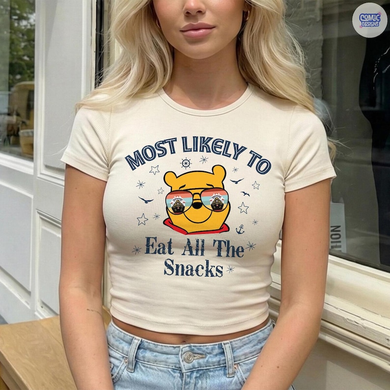May include: Cream-colored, short-sleeved crop top with the text "MOST LIKELY TO Eat All The Snacks" in blue. The graphic features a yellow bear wearing sunglasses. The shirt is ribbed and has a fitted silhouette.