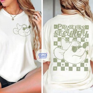 May include: Cream-colored t-shirt featuring a cartoon mouse wearing sunglasses. The front of the shirt displays the mouse graphic, while the back has the text "PRINCESS SECURITY" in a green, checkered design. The shirt is from Comic Designs.