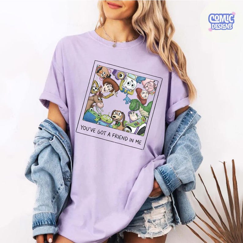 May include: A light purple t-shirt featuring a square graphic print of various animated characters arranged in a circle. The text below the graphic reads, "YOU'VE GOT A FRIEND IN ME." The shirt is worn with a blue denim jacket and denim shorts.