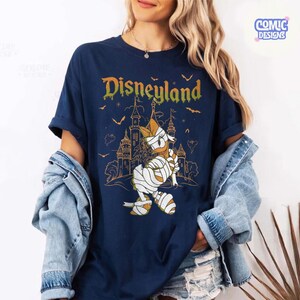 Może przedstawiać: Granatowy t-shirt z grafiką przedstawiającą postać kaczora owiniętego w bandaże na tle zarysu zamku. Napis Disneyland jest nadrukowany ż&oacute;łto-pomarańczowym gradientem nad postacią, w otoczeniu małych sylwetek nietoperzy.