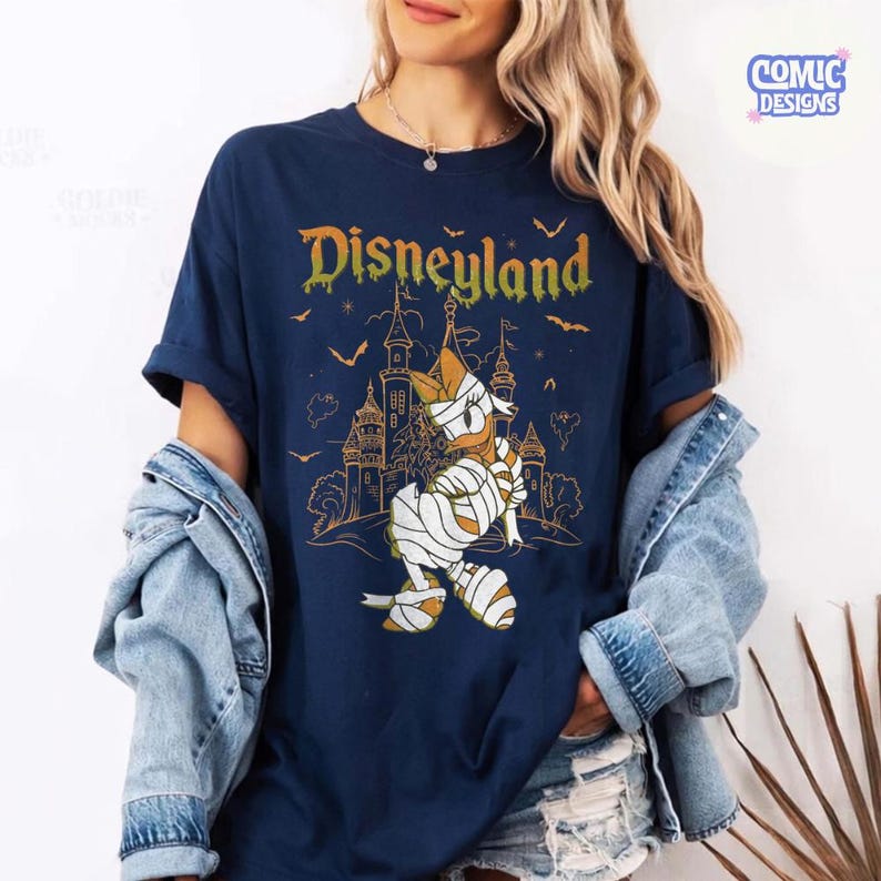 Może przedstawiać: Granatowy t-shirt z grafiką przedstawiającą postać kaczora owiniętego w bandaże na tle zarysu zamku. Napis Disneyland jest nadrukowany ż&oacute;łto-pomarańczowym gradientem nad postacią, w otoczeniu małych sylwetek nietoperzy.