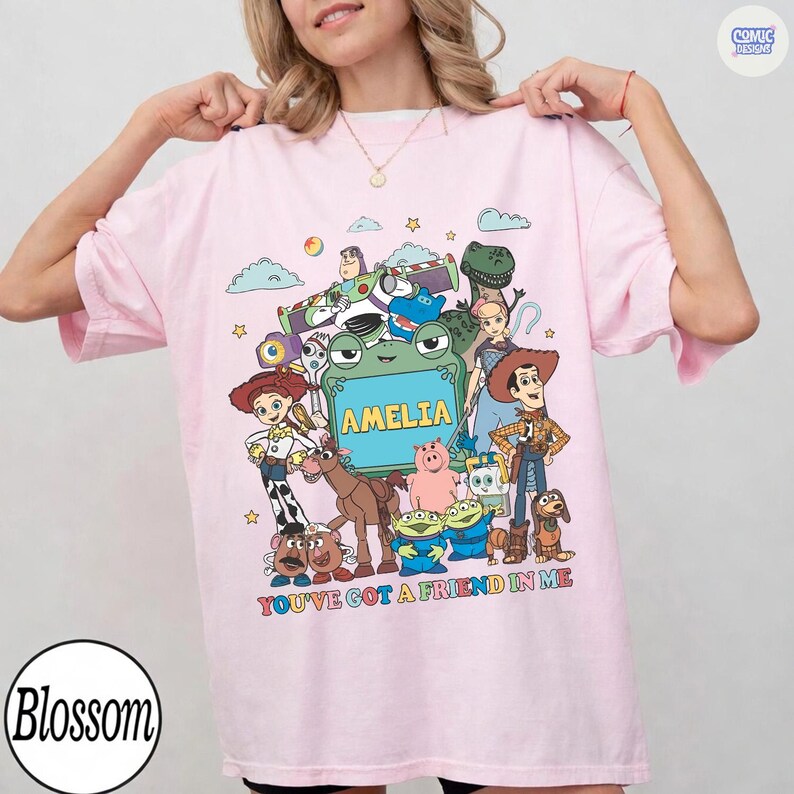 May include: A light pink t-shirt featuring a colorful graphic print of various animated characters from the Toy Story franchise. The center includes a green frog-shaped frame with the name AMELIA printed in yellow, and the text YOU'VE GOT A FRIEND IN ME.