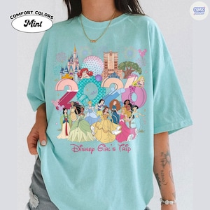 May include: A mint green t-shirt featuring a graphic print of the year 2024 with various animated princesses, castle landmarks, and the text Disney Girl's Trip. The design includes colorful illustrations of characters and park icons.