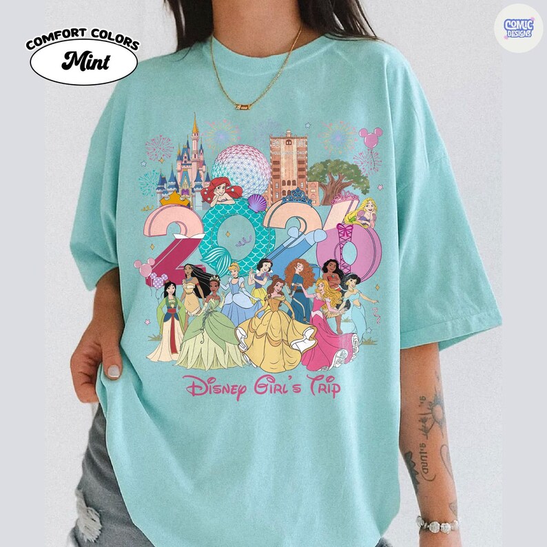 May include: A mint green t-shirt featuring a graphic print of the year 2024 with various animated princesses, castle landmarks, and the text Disney Girl's Trip. The design includes colorful illustrations of characters and park icons.