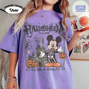 Puede incluir: Una camiseta violeta con un estampado gr&aacute;fico de un rat&oacute;n de dibujos animados con un disfraz de esqueleto frente a un castillo. El dise&ntilde;o incluye calabazas, una luna llena, telara&ntilde;as y el texto Halloween Party 2026 Not-so-scary.