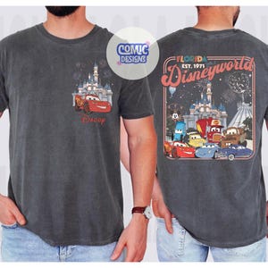 K&ouml;nnte beinhalten: Dunkelgraues T-Shirt mit einem Cars-Motiv. Auf der Vorderseite ist Lightning McQueen vor einem Schloss mit Feuerwerk und dem Wort "Daddy" zu sehen. Auf der R&uuml;ckseite befindet sich eine Disney World-Grafik mit Cars-Charakteren und dem Text "Florida Est. 1971".