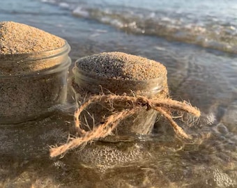 Può includere: Due barattoli di vetro pieni di sabbia, uno legato con uno spago, seduti su una spiaggia con onde sullo sfondo.