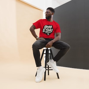 May include: A red t-shirt with the text "OMO ESHU" in a black and white design. The person is wearing black jeans and white sneakers. The background is a mix of beige and black.