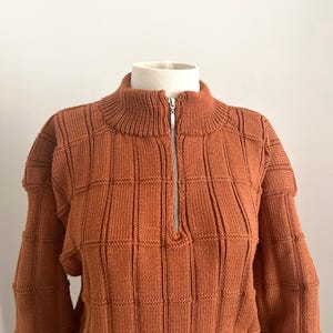 Puede incluir: Un suéter de color marrón anaranjado con cuello alto y cremallera plateada. El suéter tiene un patrón texturizado en forma de cuadrícula. La prenda se exhibe sobre una cabeza y hombros de maniquí blancos.