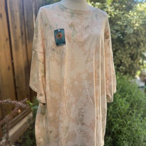 May include: Oversized, tie-dye t-shirt in shades of beige and green, featuring a rectangular patch with a sun design and the text "The Sun". The shirt is displayed on a mannequin, showcasing its loose fit and casual style.