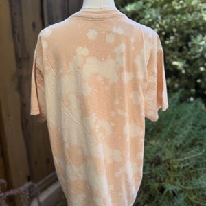 May include: A peach-colored t-shirt with a bleached, spotted pattern. The shirt is displayed on a mannequin, showcasing the back of the garment. The fabric appears soft and casual, suitable for everyday wear.