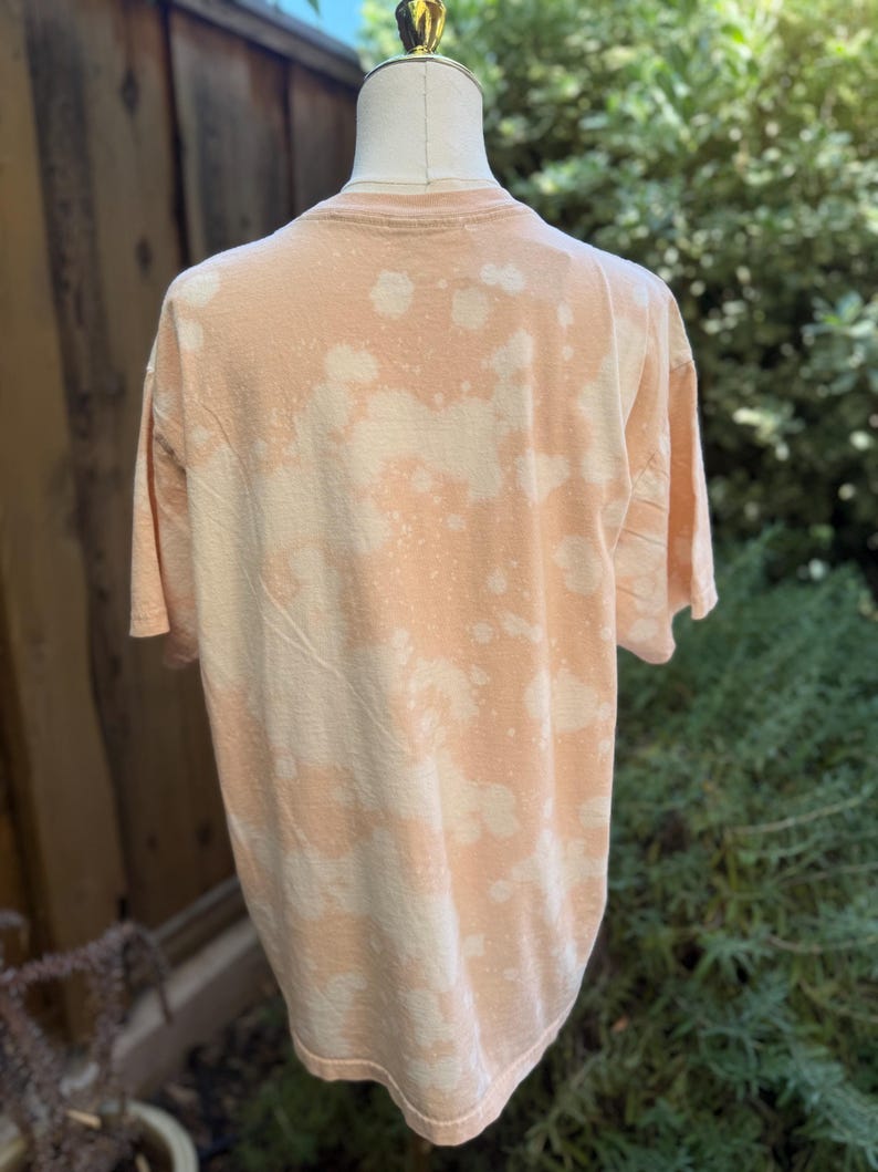 May include: A peach-colored t-shirt with a bleached, spotted pattern. The shirt is displayed on a mannequin, showcasing the back of the garment. The fabric appears soft and casual, suitable for everyday wear.