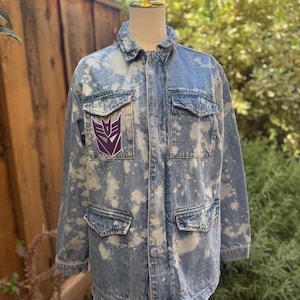 May include: A light blue denim jacket with a bleach-splatter effect. The jacket features four front pockets and a purple Transformers logo patch on the chest. The jacket is displayed on a mannequin.